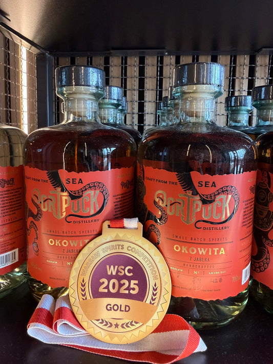 Złoty medal Warsaw Spirits Competition WSC 2025 leżący przed butelkami rzemieślniczej Okowity z Jabłka od Port Puck Distillery.