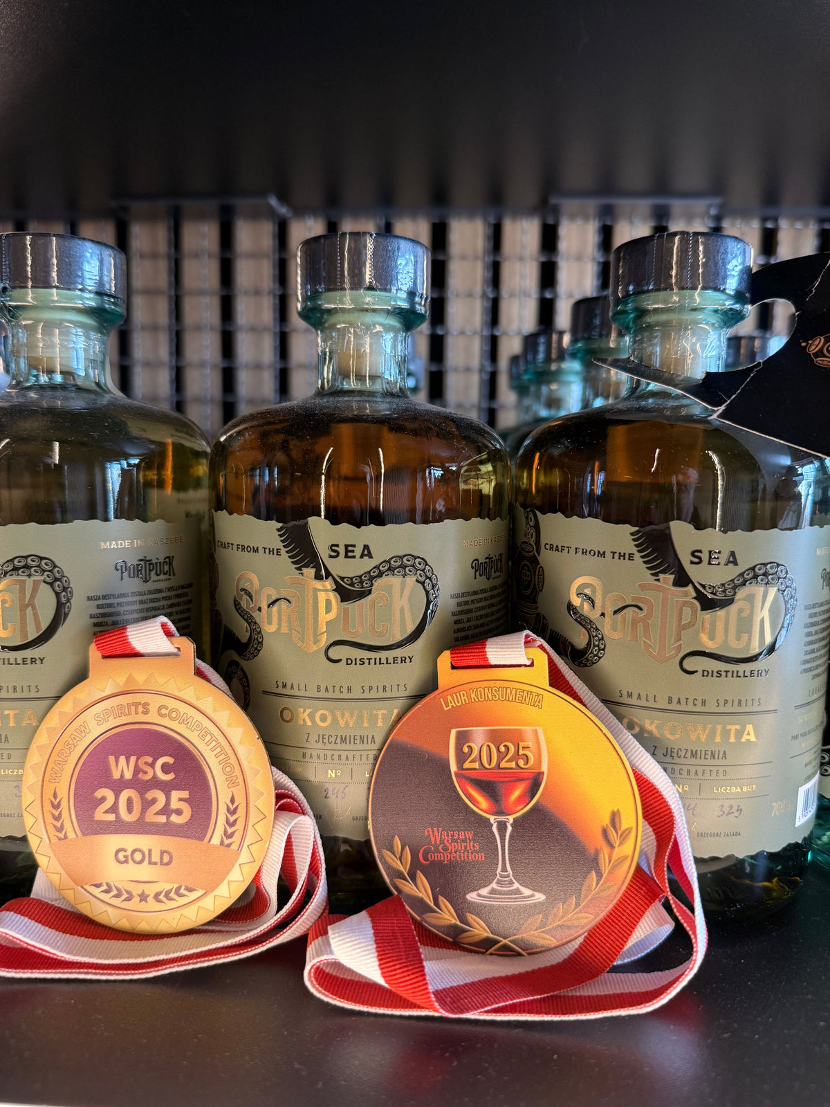 Dwa złote medale Warsaw Spirits Competition WSC 2025 zawieszone na butelkach rzemieślniczej Okowity Jęczmiennej z Port Puck Distillery.