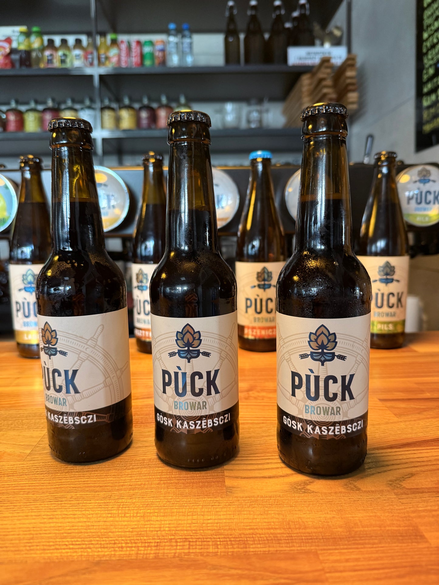 Trzy butelki piwa Gòsk Kaszëbsczi Russian Imperial Stout stojące na drewnianym barze w Browarze Pùck