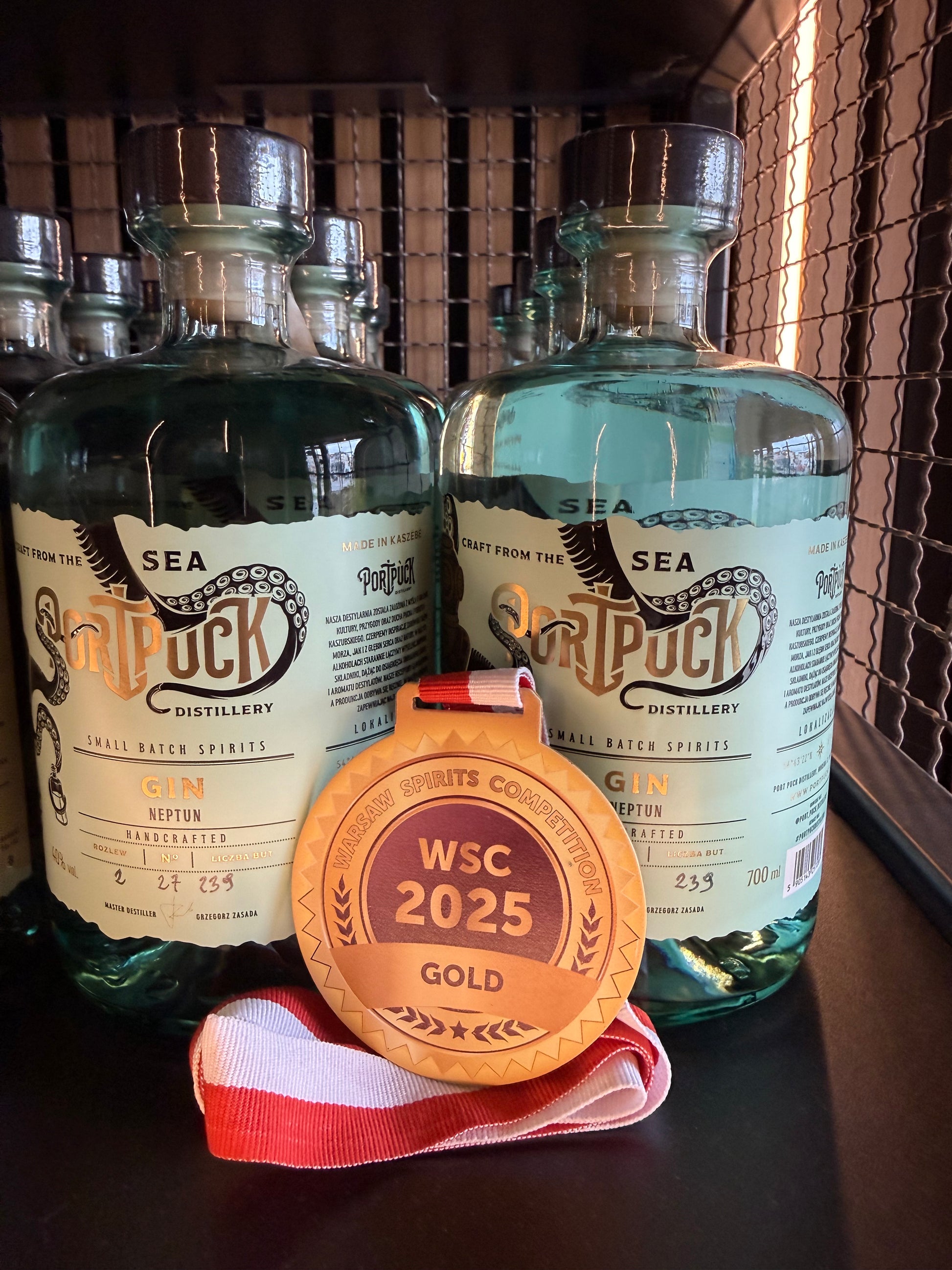 Złoty medal Warsaw Spirits Competition WSC 2025 zaprezentowany na tle butelek rzemieślniczego Ginu Neptun z Port Puck Distillery.
