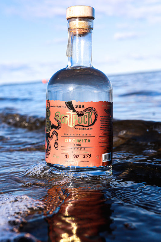 Butelka rzemieślniczej Okowity z Piwa z Port Puck Distillery stojąca w morskich falach na brzegu Zatoki Puckiej.
