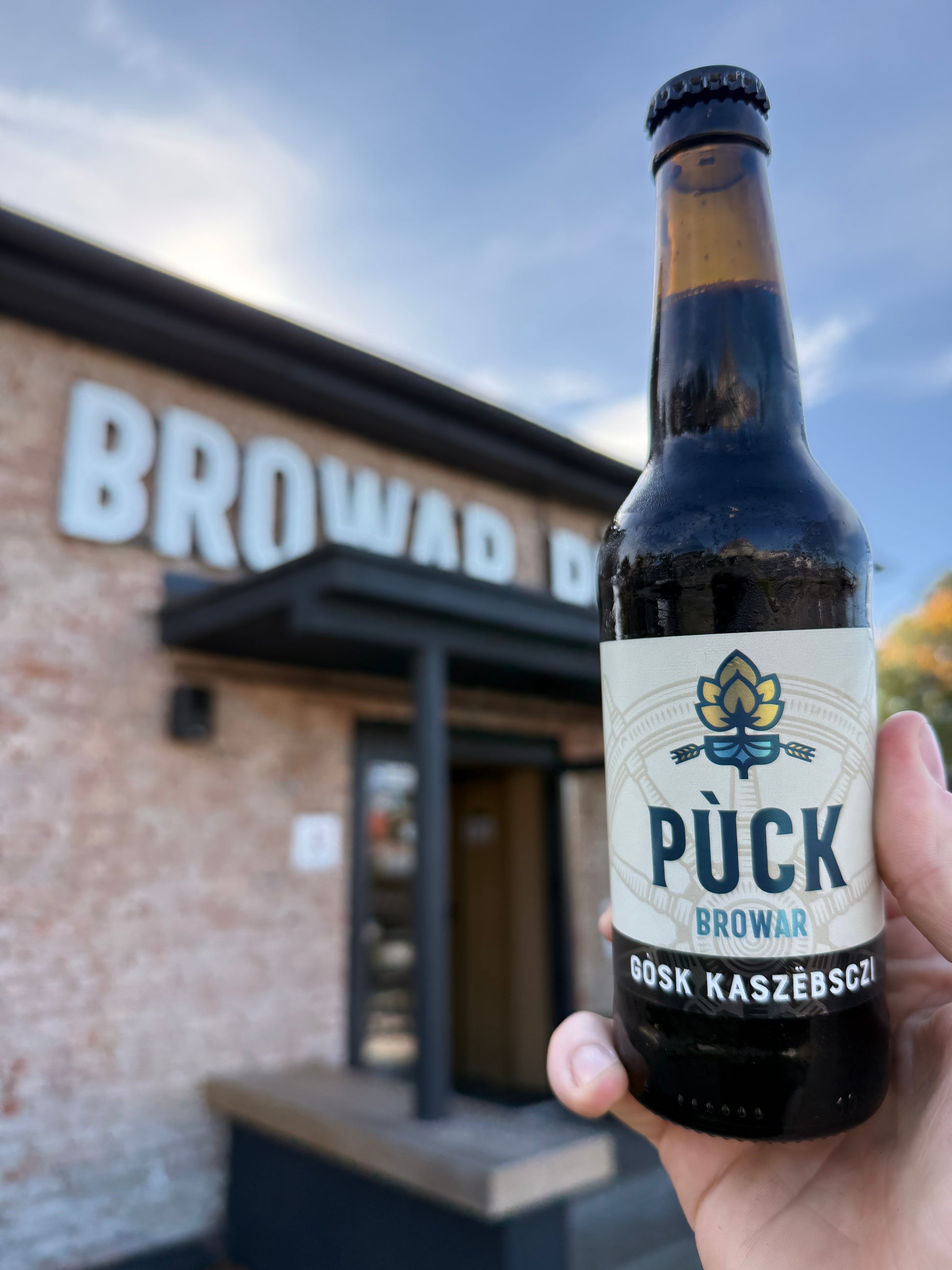 Butelka piwa Gòsk Kaszëbsczi Russian Imperial Stout 10,5% trzymana w dłoni na tle budynku Browaru Pùck