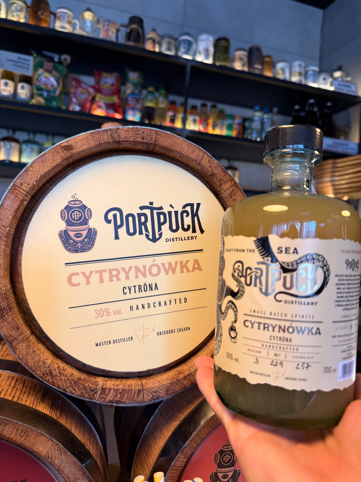 Butelka rzemieślniczej Cytrynówki trzymana w dłoni obok drewnianej beczki z logo Port Puck Distillery w klimatycznym barze.
