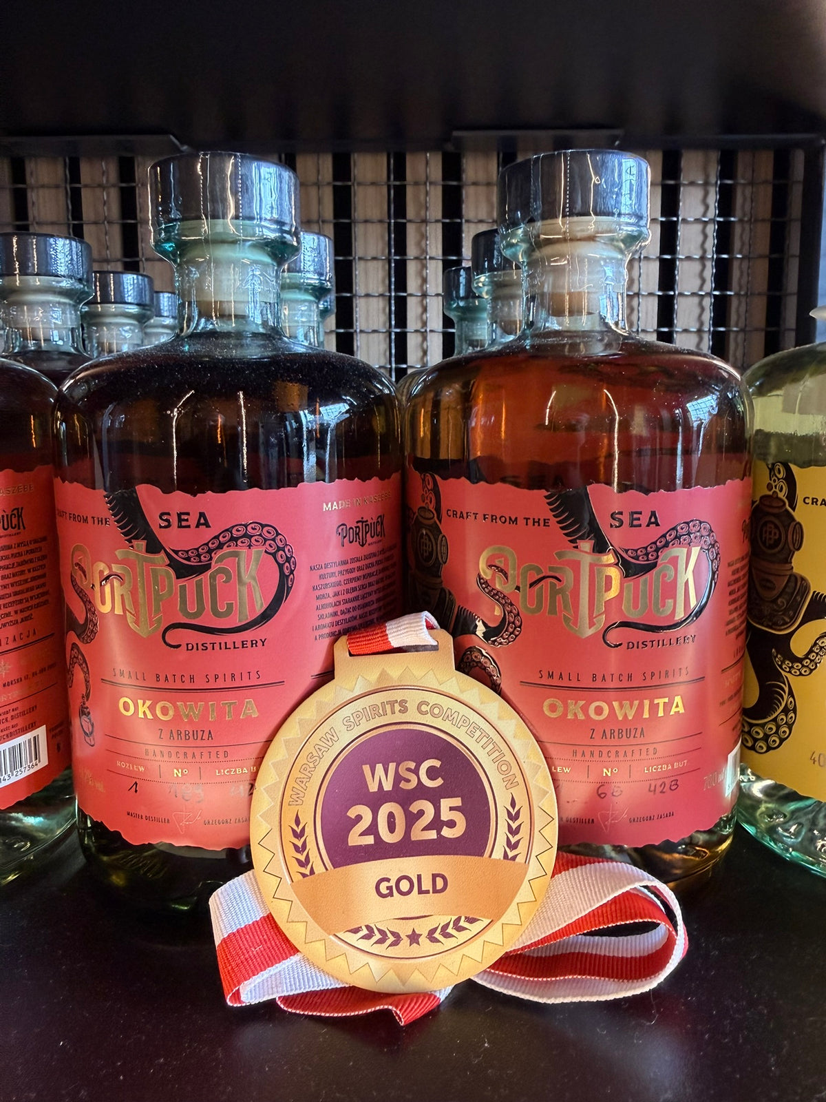 Złoty medal Warsaw Spirits Competition WSC 2025 zaprezentowany przed butelkami rzemieślniczej Okowity z Arbuza z Port Puck Distillery.
